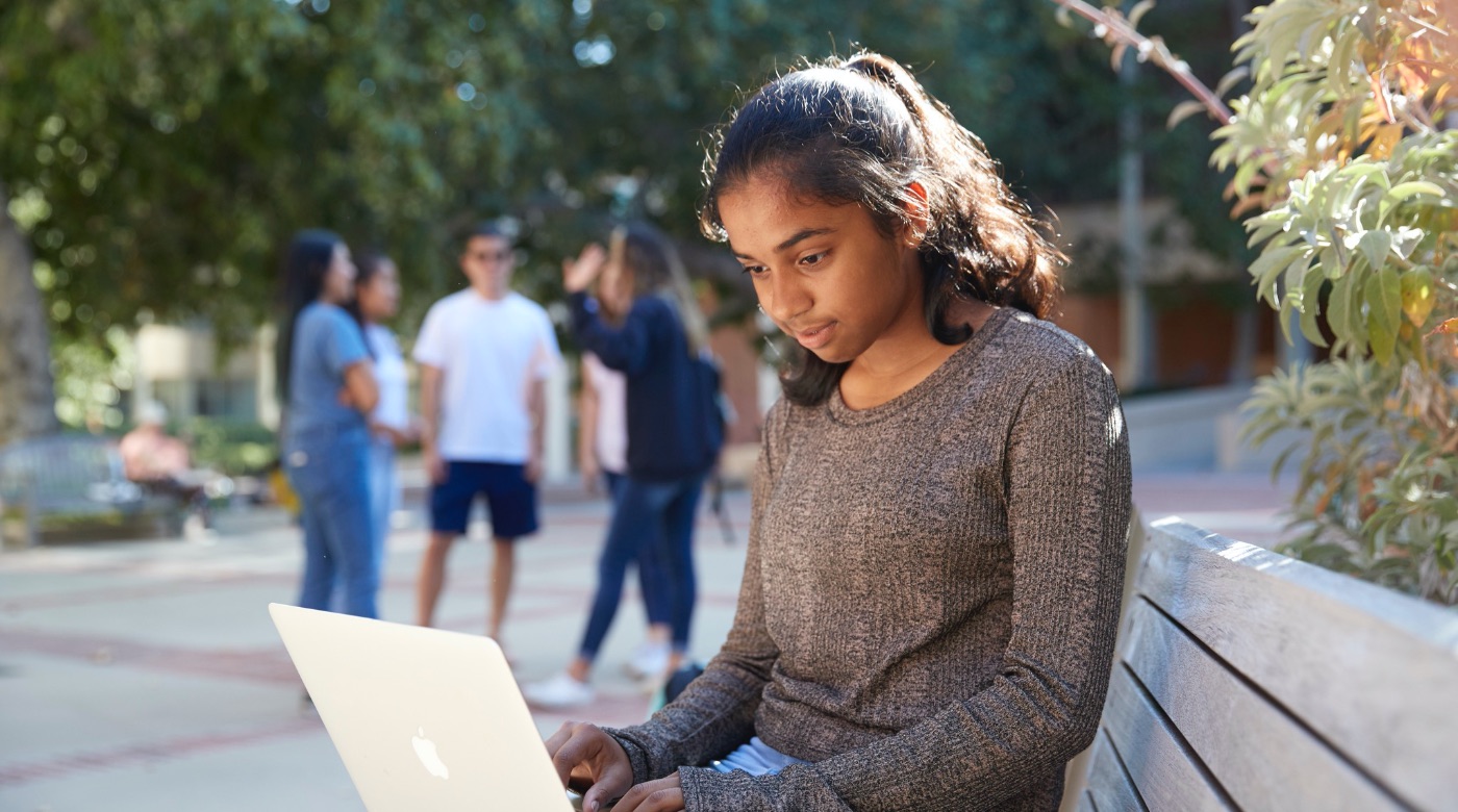 Student working on her laptop on a bench as a group of students behind her stand and talk.