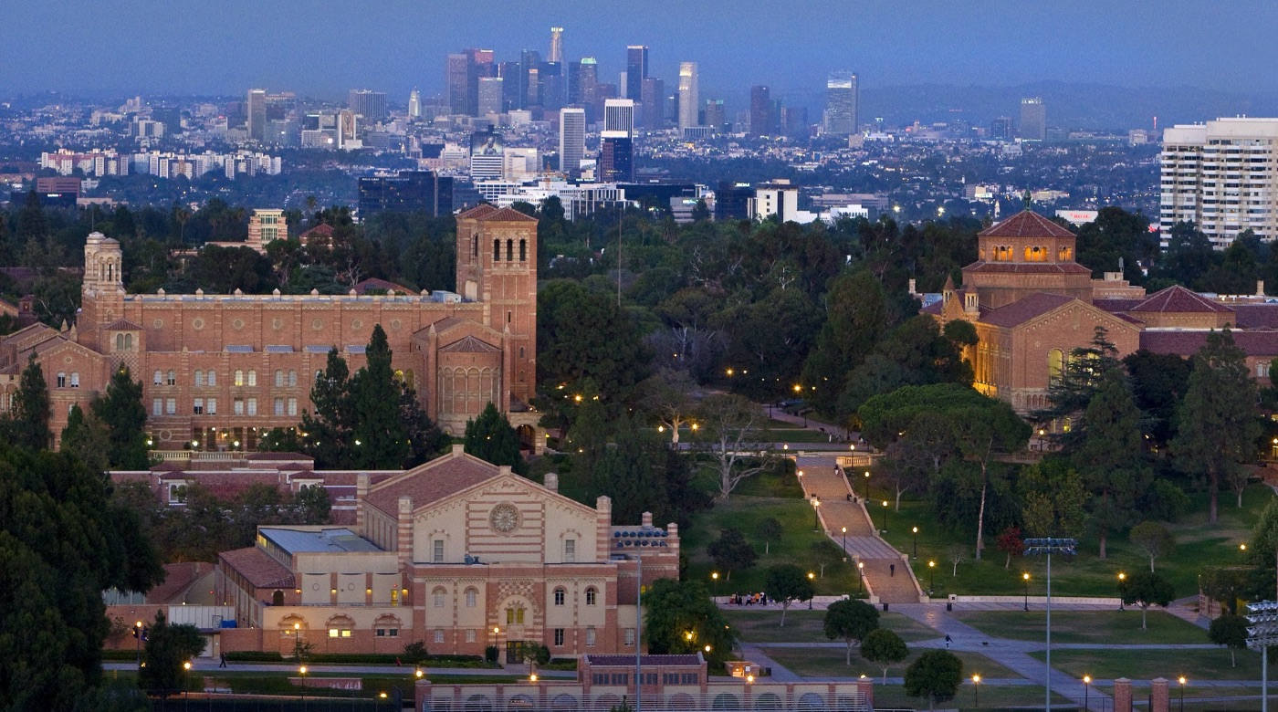 A panoramic view of Powell Library, Janss Steps, and Royce Hall at night. In the background, a view of Downtown Los Angeles is visible.