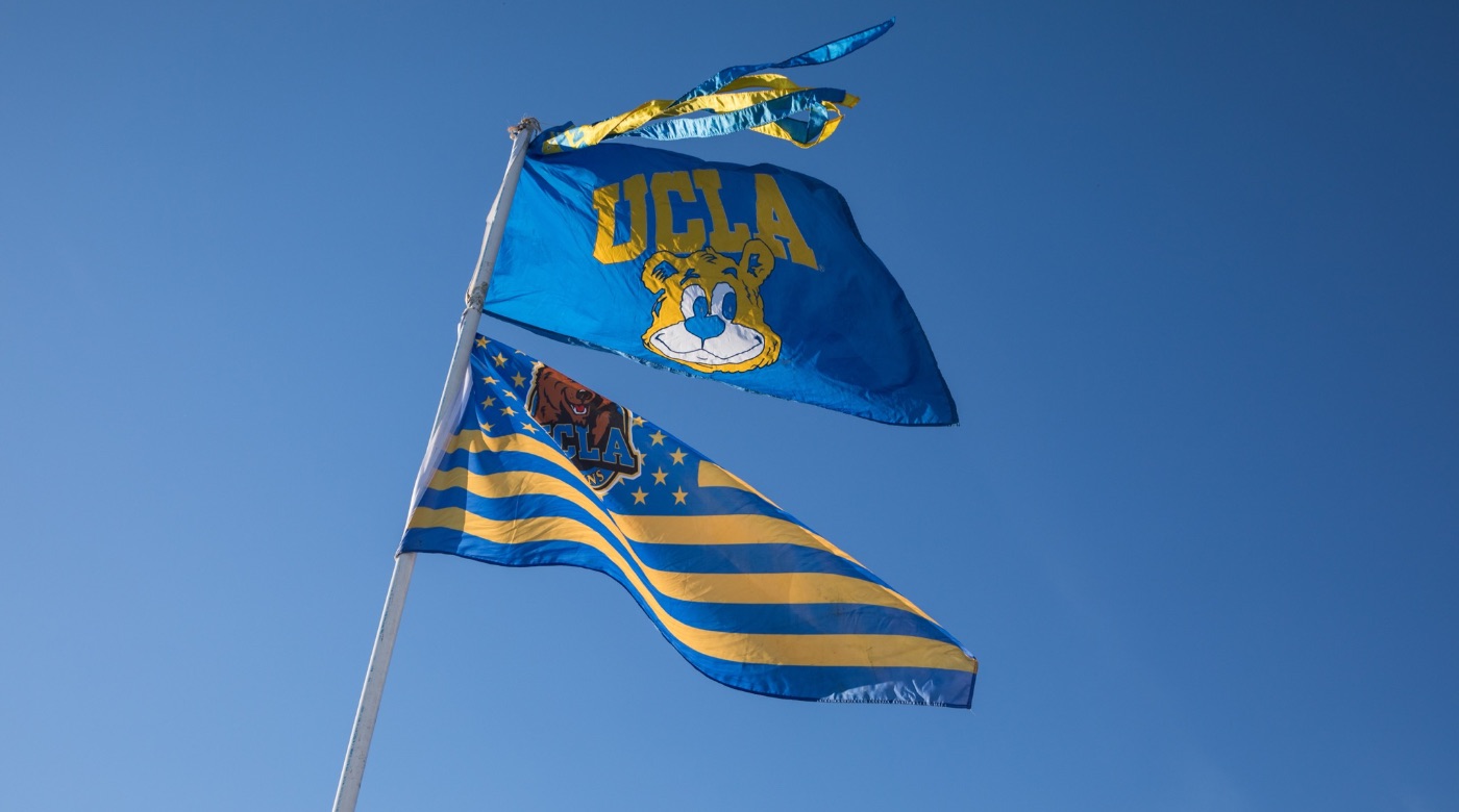 Three UCLA flags hoisted on a pole. The first flag is alternating blue and yellow stripes, the second flag has a blue background with UCLA and the Bruin bear in yellow, the third flag is the U.S. flag with a Bruin bear in the middle of yellow stars with alternating blue and yellow stripes.