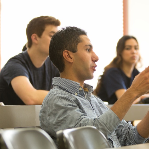 Student answering question in a classroom. Two students behind him are listening to his answer.