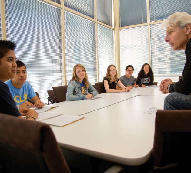 A professor speaking with a student as five other students watch them. All students are seated at a table while the professor is directly looking at one student.