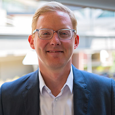 Photograph of Adam Steig. He is wearing a blue coat, light blue shirt and glasses.