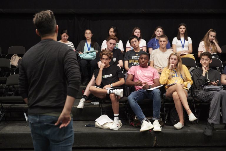 Three rows of students take notes and listen to an instructor standing in front of them with his back to the camera in a dark performance studio.