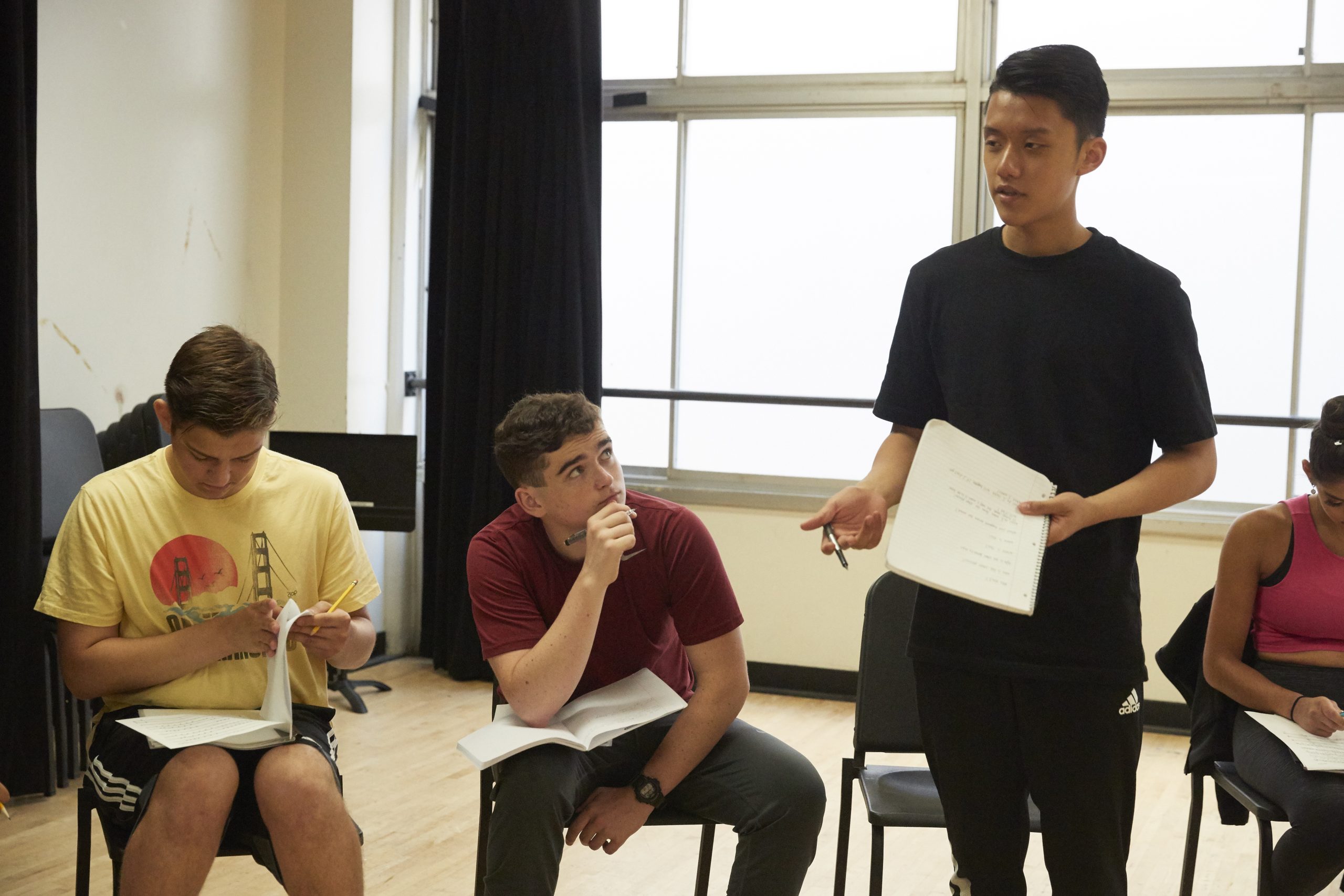 A male student stands in a classroom holding a notebook and pen with three students sitting next to him with notebooks on their laps, one looking up at him.