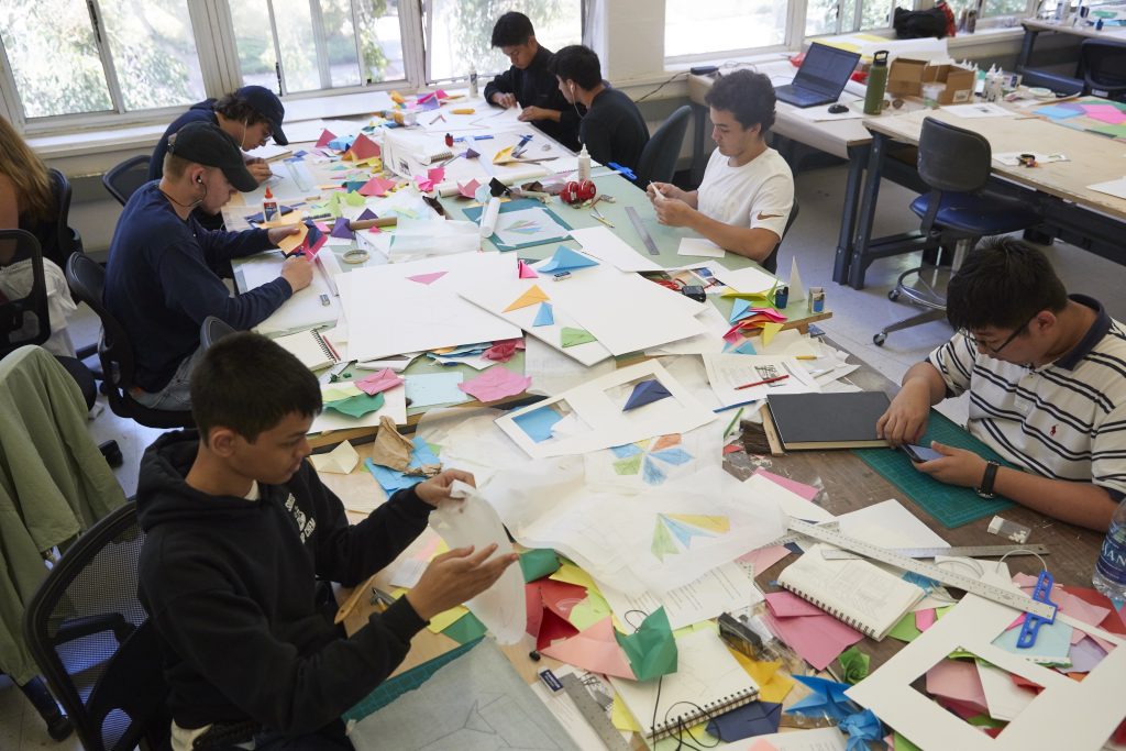 Overhead view of seven students working at a large table covered with colored paper, notebooks, rulers and other art supplies