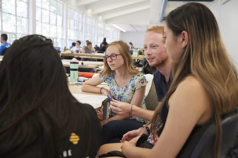 Three female students and one male student sit together at a table talking in a classroom, with other students in the background.