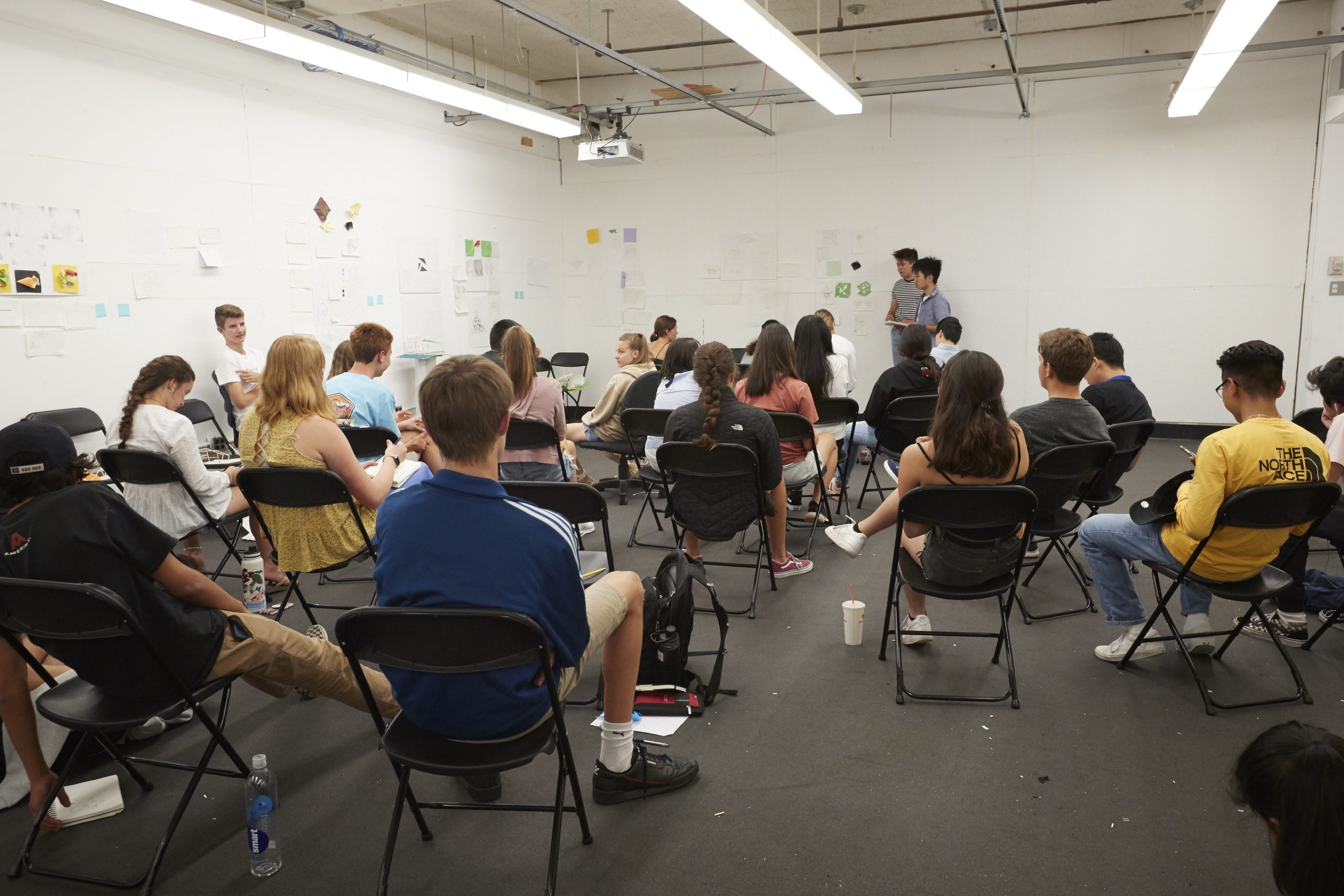 Students sit in folding chairs in a room with white walls and student artwork hung on the walls, facing two students who are standing in the front of the room.