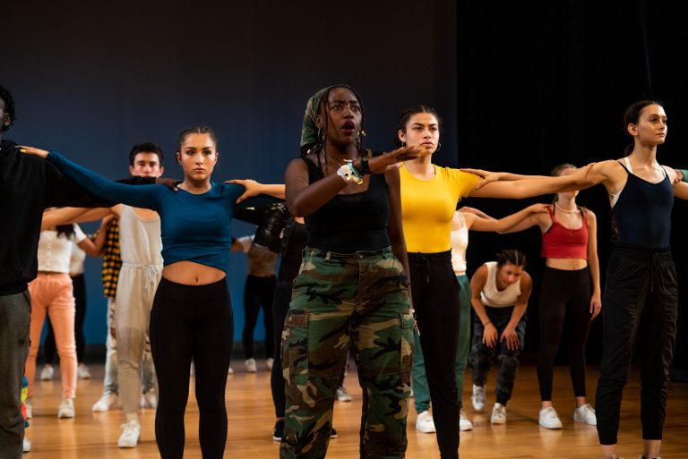 Students standing close together with arms outstretched, doing movement exercises in a dance studio.