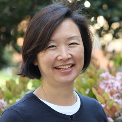 Portrait of Dr. Jennifer Jung-Kim. She is wearing a blue sweater and a white shirt. She has chin length dark brown hair and a bright smile.