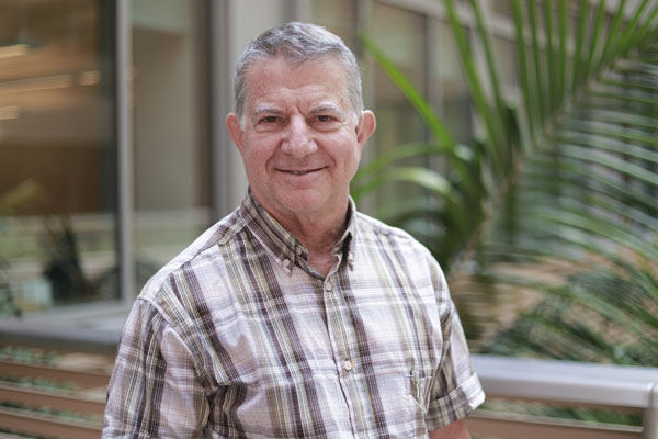 Portrait of Eli Gafni. He is wearing a casual grey plaid shirt and standing in front of a palm tree.