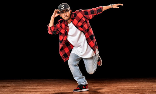 Portrait of Ernesto Galarza. He is wearing light blue denim jeans, a red plaid shirt opened showing the white t-shirt beneath, a black baseball cap, red and black sneakers and is standing in a hip hop dance move.