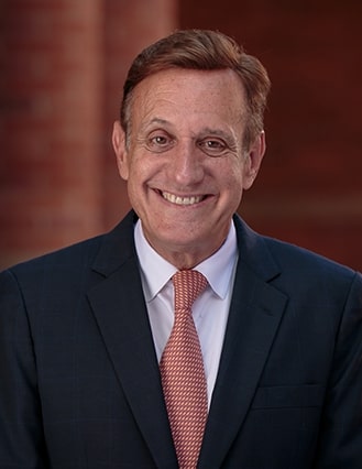 Portrait of Gonzalo Freixes. He is wearing a dark suit jacket, a white shirt and brown tie.