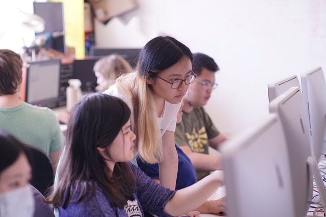 A female student stands behind a female student at a computer in a computer lab, examining her screen.