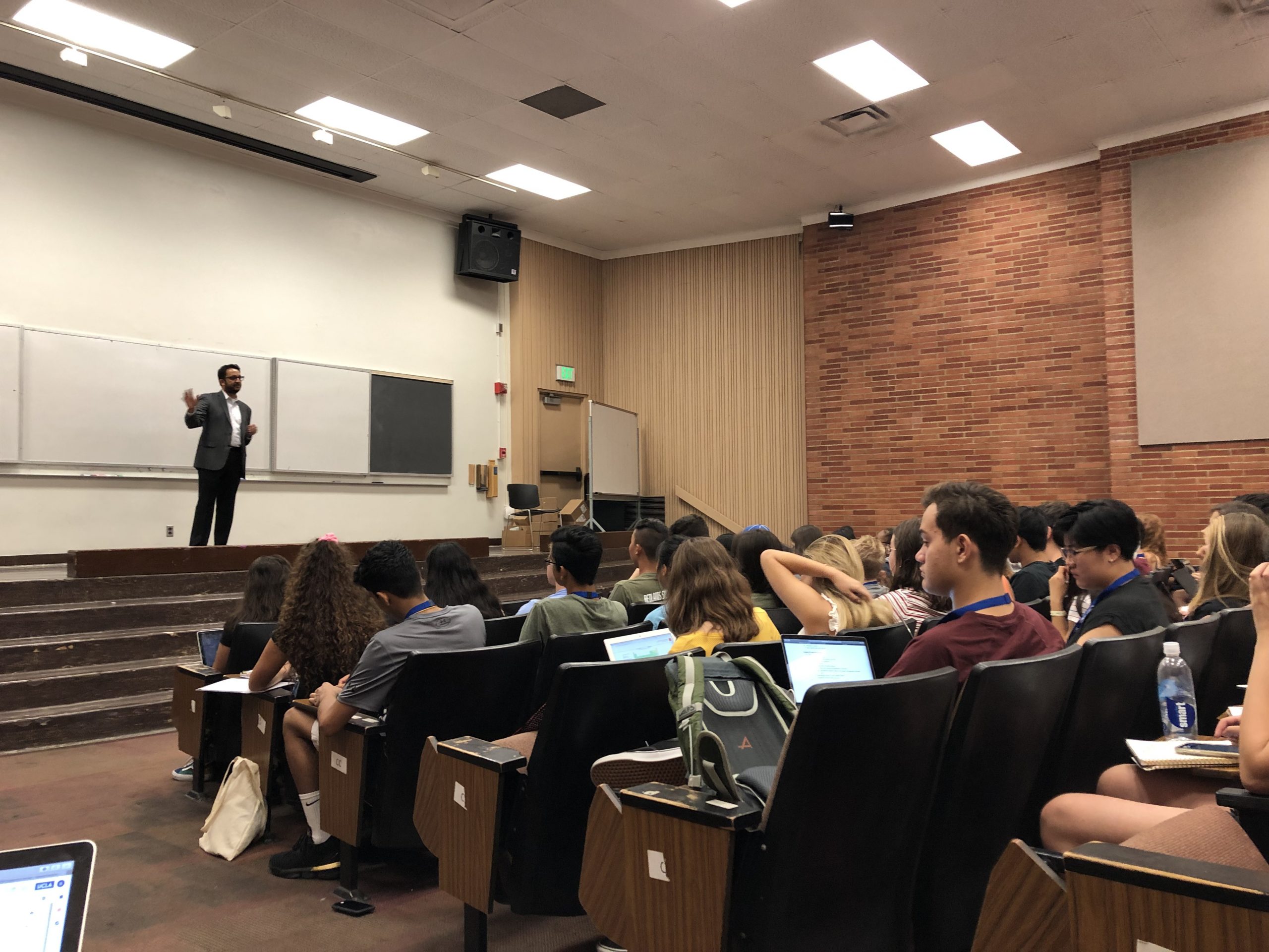 An instructor stands in front of a whiteboard in the front of a lecture hall full of students.