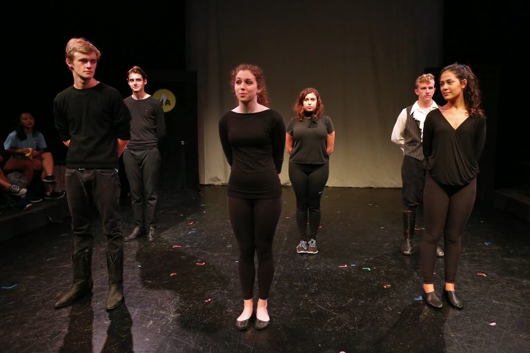Six students dressed in black stand in a dim performance studio with a black floor.
