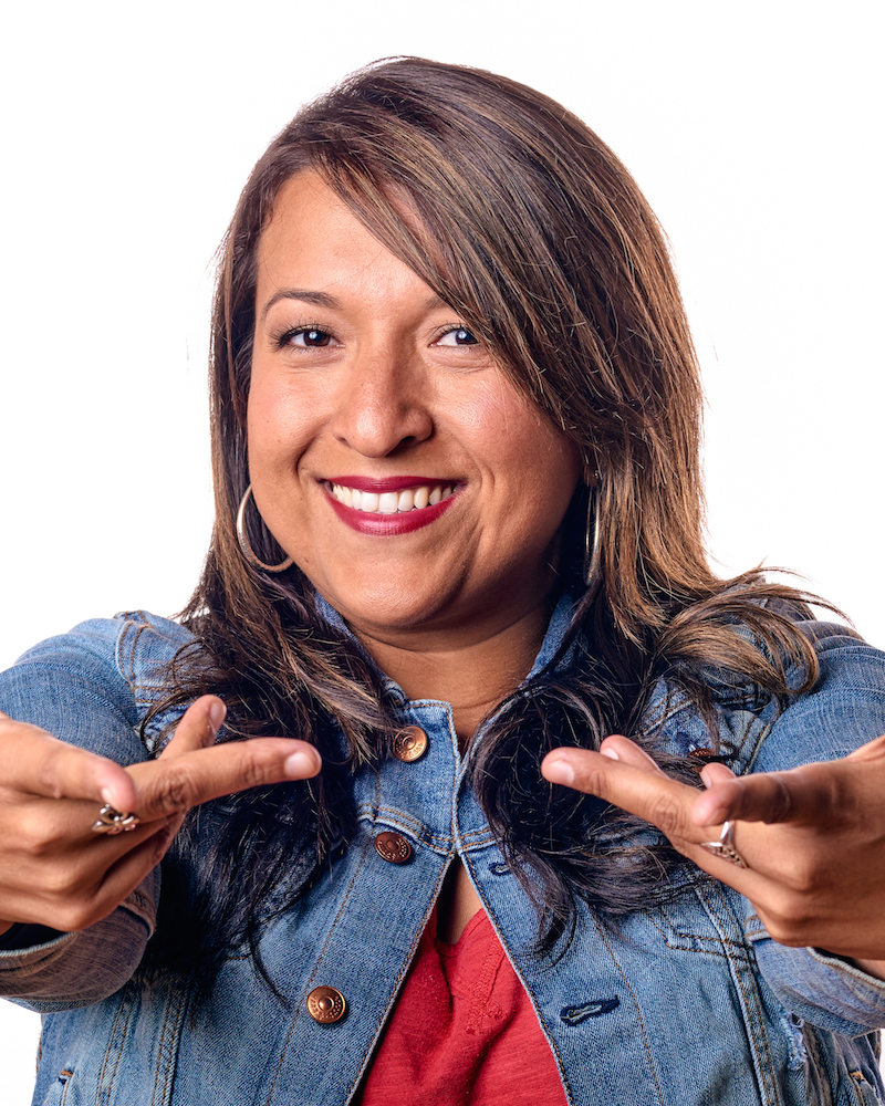 Portrait of Jackie Lopez. She is wearing a denim light blue jacket, red shirt, brown long hair and has her hands stretched in front of her towards the camera.