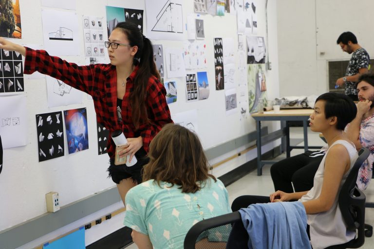 A standing female UCLA Introduction to Architecture student points to a wall of student work while three seated classmates look on.