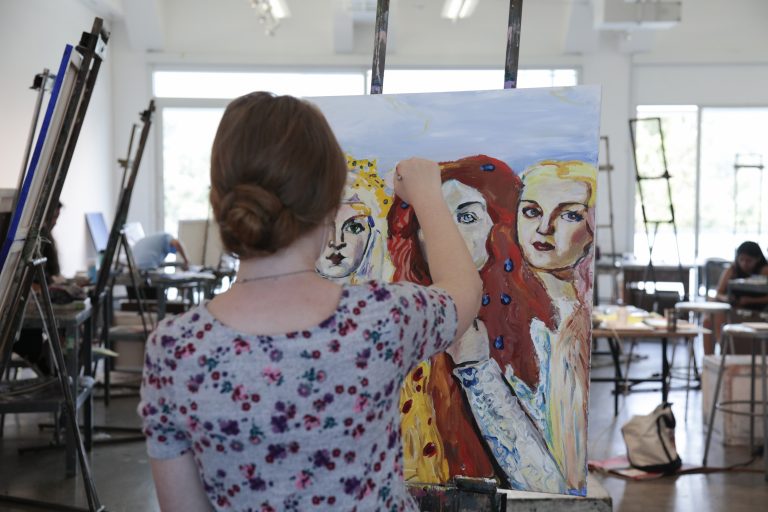 A female student in an art studio holds a paintbrush to a painting of three women.