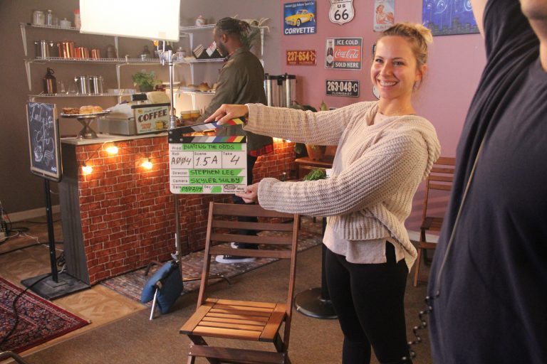 A smiling female student holds up a clapperboard in a coffee shop with lighting equipment in the background.