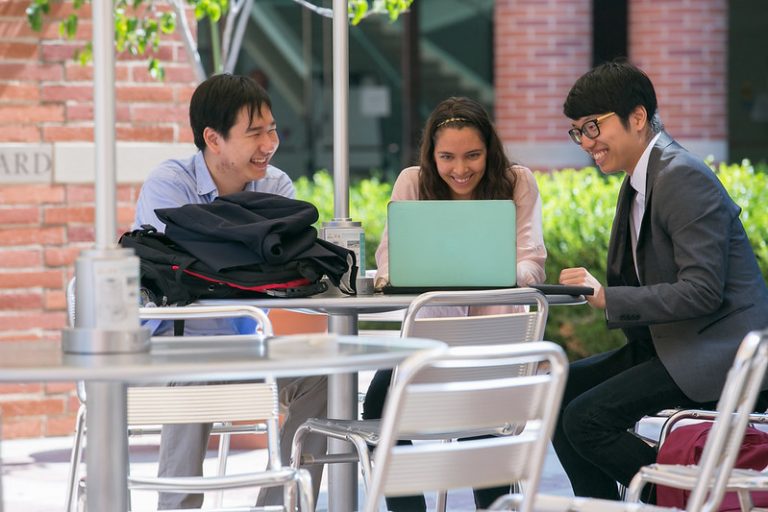 Three UCLA students smile while sitting at an outdoor table. There are two males and one female. Both males look on to the laptop of the female student.