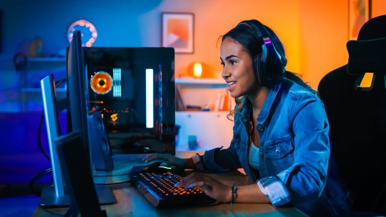 Smiling female UCLA student plays video game on colorfully lit computer in her dorm room.
