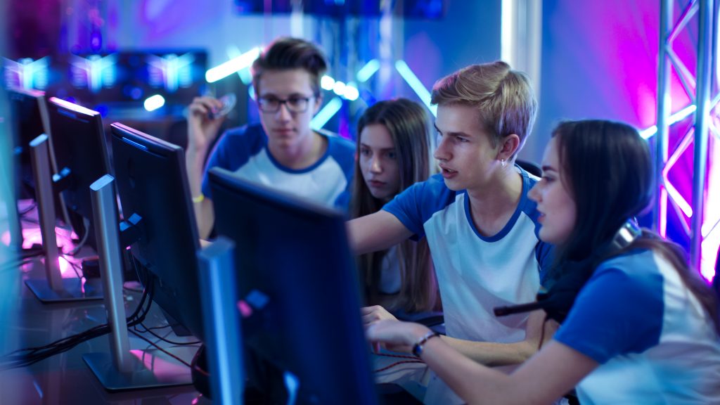 Four teenagers wearing matching white and blue T-shirts look at a computer, with blue, purple and pink lighting around them.