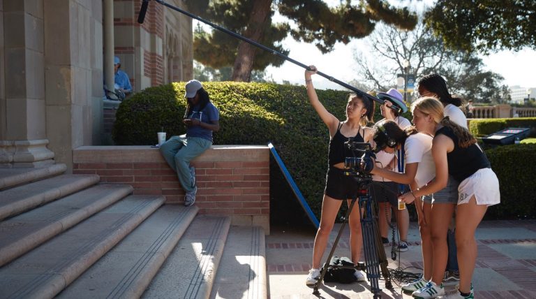 Group of students filming at the bottom of steps while another student is sitting on the ledge of the steps.