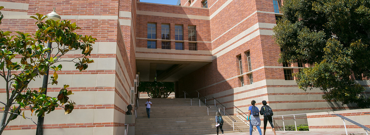 A large red brick academic building with concrete stairs going through the middle and students walking by.