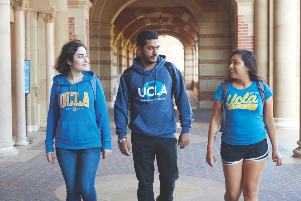 Three students walk together through a university corridor, wearing UCLA hoodies and t-shirts, smiling and engaging with each other.