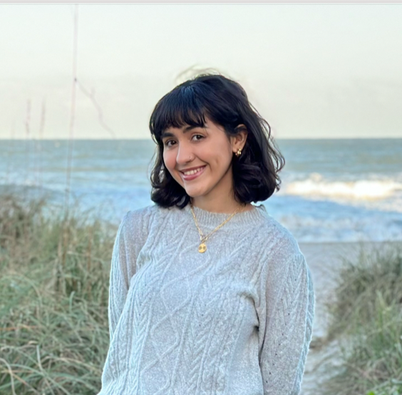 Young woman with dark, curly hair smiles at the camera, wearing a light sweater, with a beach scene and ocean in the background.