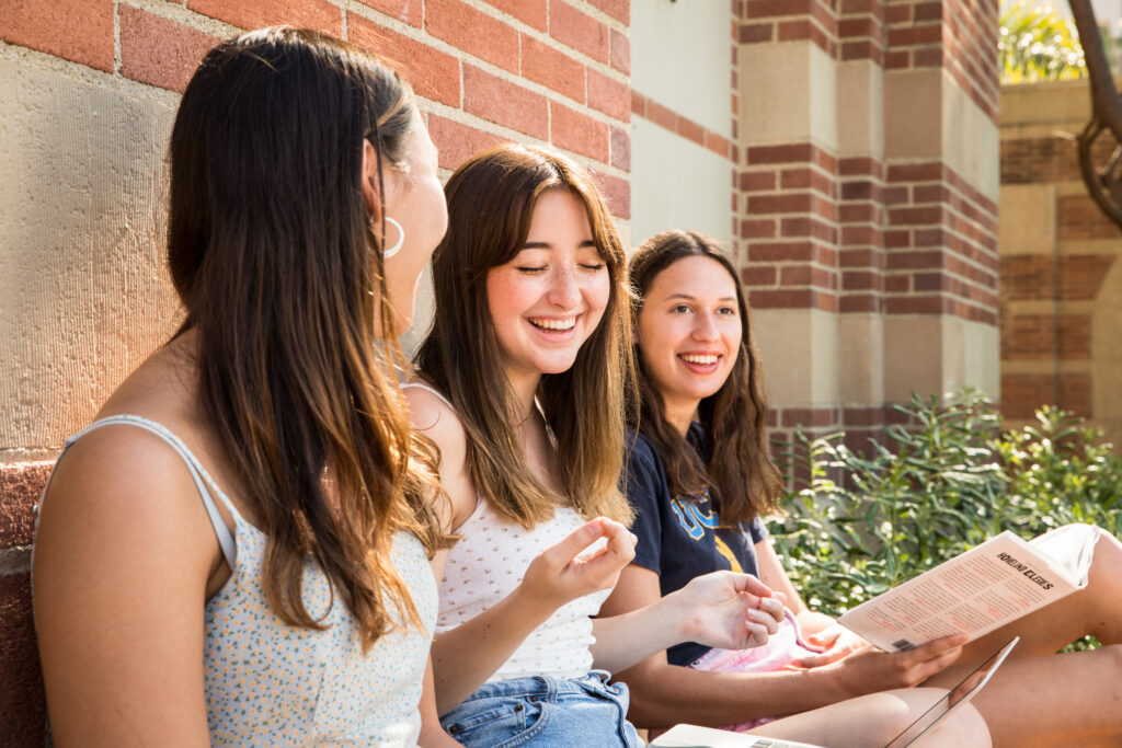 Three smiling young women sitting together on a brick wall, engaged in conversation while holding papers, surrounded by greenery.