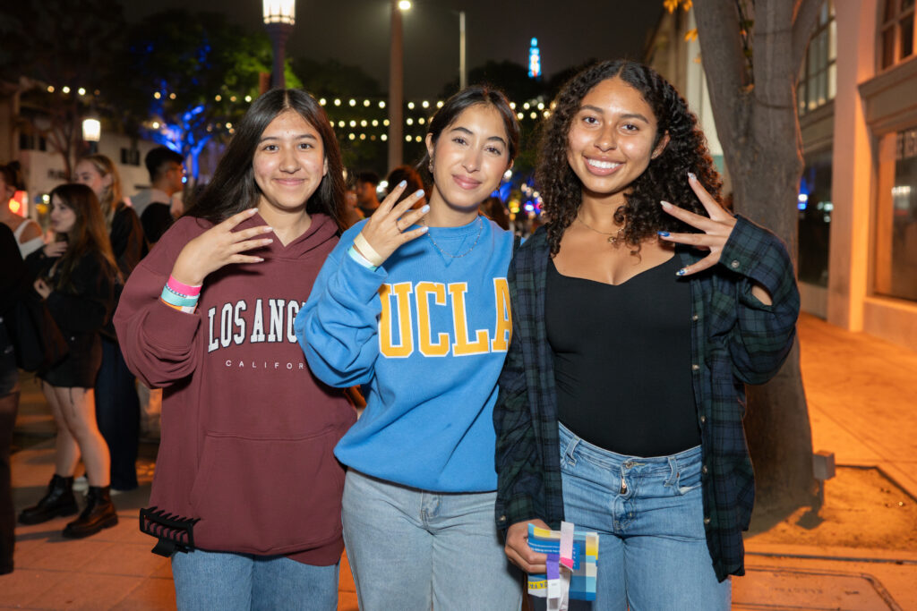 Three young women smile and pose together outdoors at night, wearing UCLA apparel and casual clothes, with festive lights in the background.
