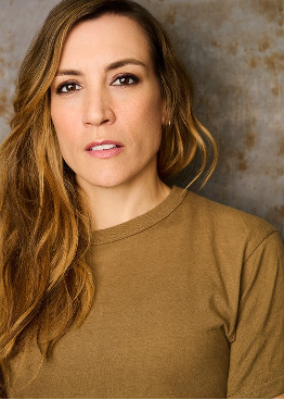 A woman with long, wavy brown hair wearing a tan shirt, poses with a neutral expression against a textured, muted background.